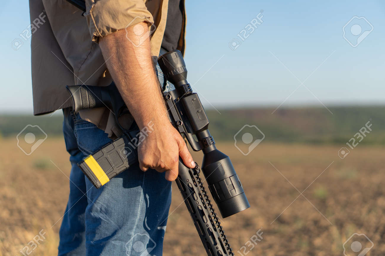 A rifle in a mans hand, close-up. Hunter with a weapon on the hunt against the background of a beautiful summer landscape - 155623098 A rifle in a mans hand, close-up. Hunter with a weapon on the hunt against the background of a beautiful summer landscape - 155623098
