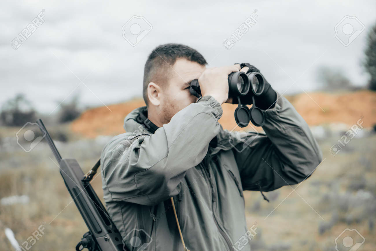 Man with a rifle looking through binoculars. Private military contractor or hunter - 164160324 Man with a rifle looking through binoculars. Private military contractor or hunter - 164160324