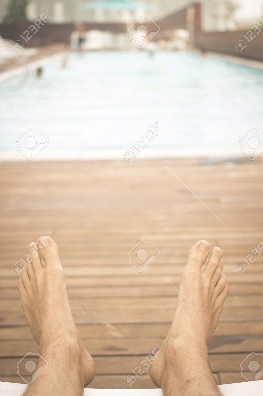 Legs And Feet Of Man Sunbathing On Sun Lounger In Summer By Swimming Pool Wooden Legs And Feet Of Man Sunbathing On Sun Lounger In Summer By Swimming Pool Wooden