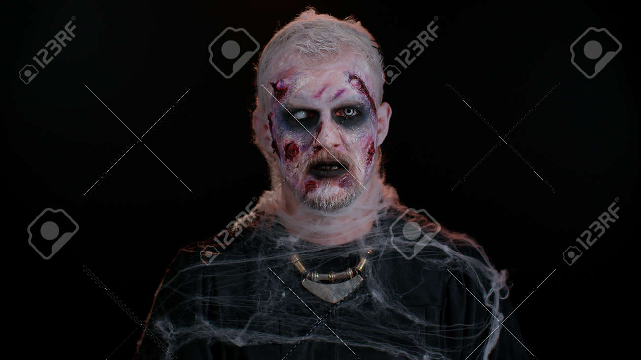 가짜 상처 흉터와 흰색 콘택트 렌즈로 화장을 한 좀비 남자는 카메라를 보고 이빨을 클릭하며 어두운 방에서 겁을 주려고 합니다. 불길한  죽은 사람. 할로윈, 촬영, 준비 개념 로열티 무료 사진, 그림, 이미지 그리고