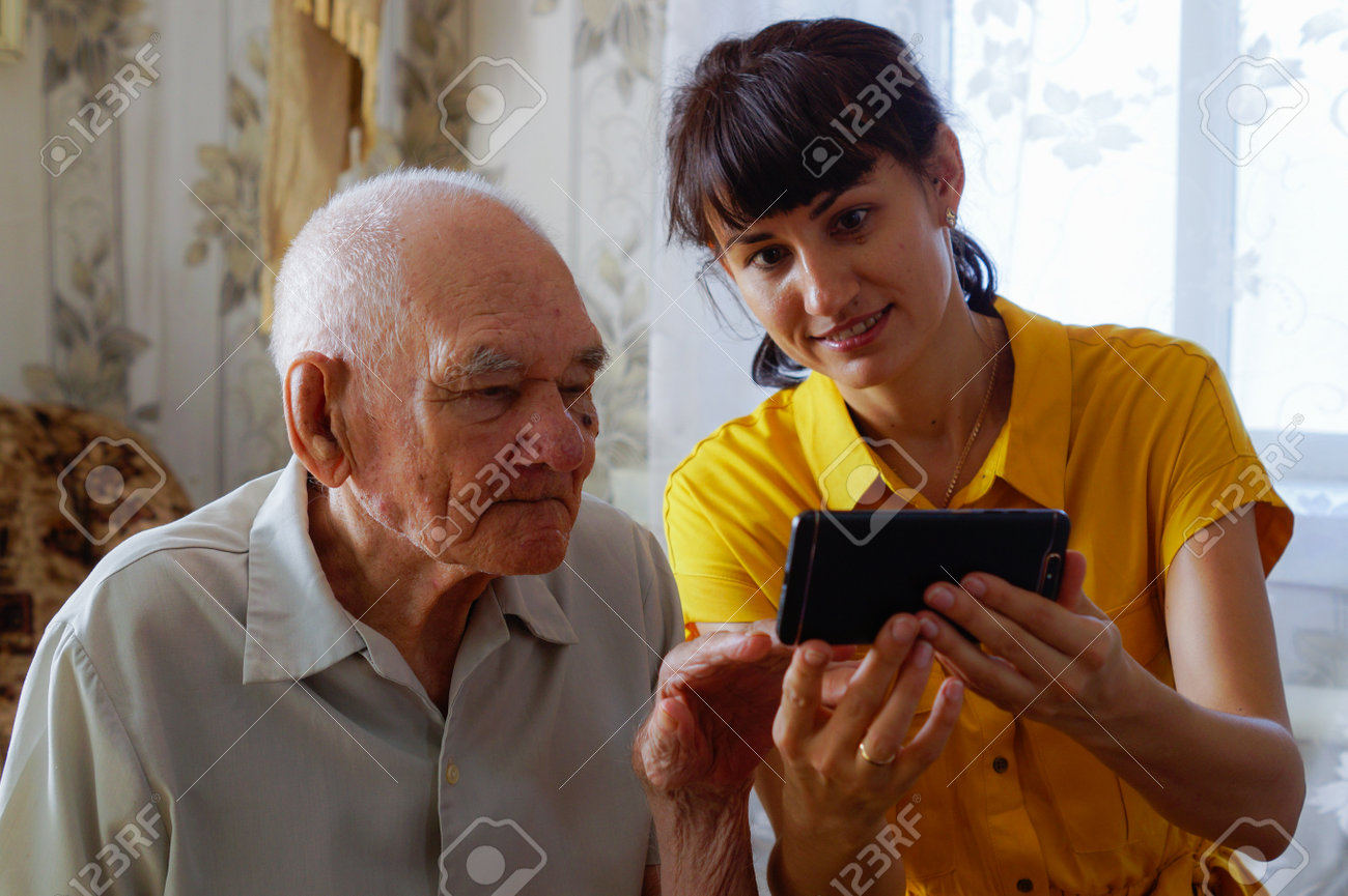 Um homem maduro de aparência europeia com uma jovem mulher sentada no sofá e olhando para o telefone. pagar por compras através do seu telefone. gerações. aprendendo tecnologia da internet on-line ou wi-fi no conceito de uma pessoa mais velha. - 162963478 Um homem maduro de aparência europeia com uma jovem mulher sentada no sofá e olhando para o telefone. pagar por compras através do seu telefone. gerações. aprendendo tecnologia da internet on-line ou wi-fi no conceito de uma pessoa mais velha. - 162963478