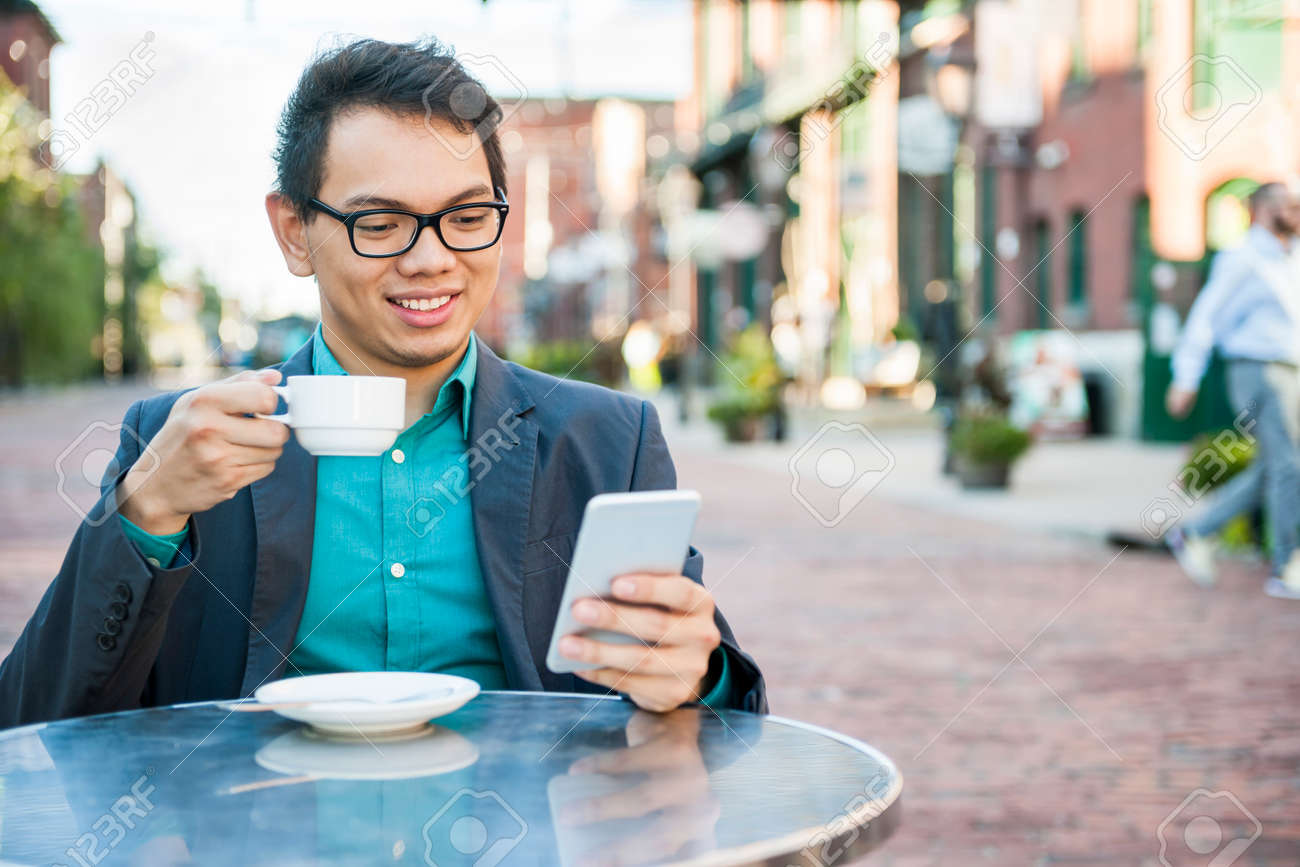 Young asian man in business casual attire sitting and smiling in relaxing outdoor cafe drinking cup of coffee while using mobile phone - 55759726 Young asian man in business casual attire sitting and smiling in relaxing outdoor cafe drinking cup of coffee while using mobile phone - 55759726