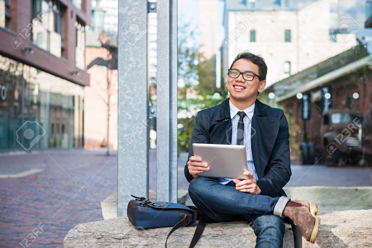Young smiling asian business man holding a digital tablet sitting outside on city street looking away - 58163849 Young smiling asian business man holding a digital tablet sitting outside on city street looking away - 58163849
