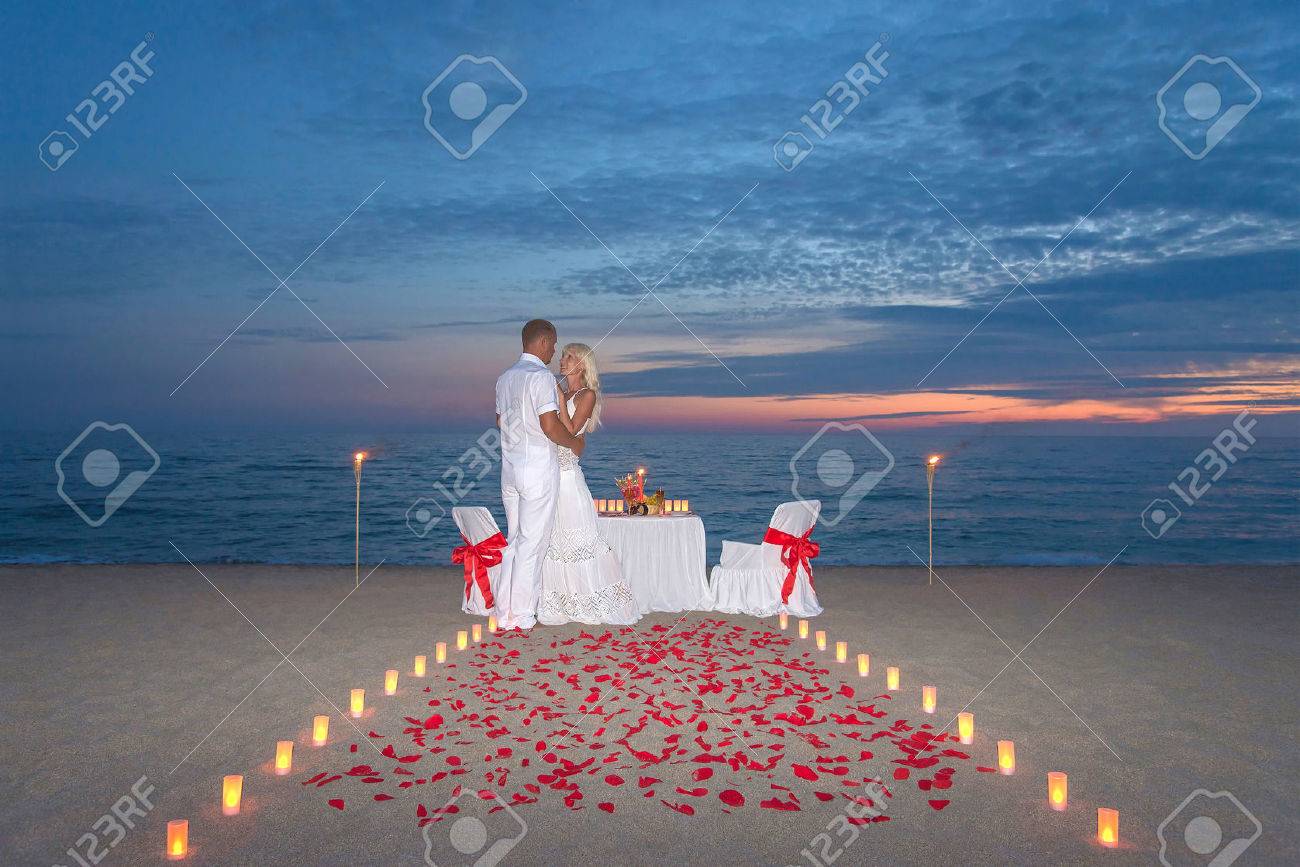 Jeune Couple Part Un Dîner Romantique Avec Des Bougies Torches Et Manière Ou Pétales De Rose Sur La Plage De Sable De Mer Contre Le Coucher Du Soleil