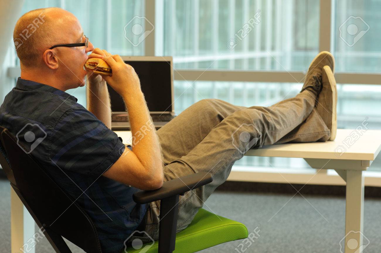 middle aged caucasian man sitting in relaxed position at the desk, eating hamburger in his office - 32612694 middle aged caucasian man sitting in relaxed position at the desk, eating hamburger in his office - 32612694