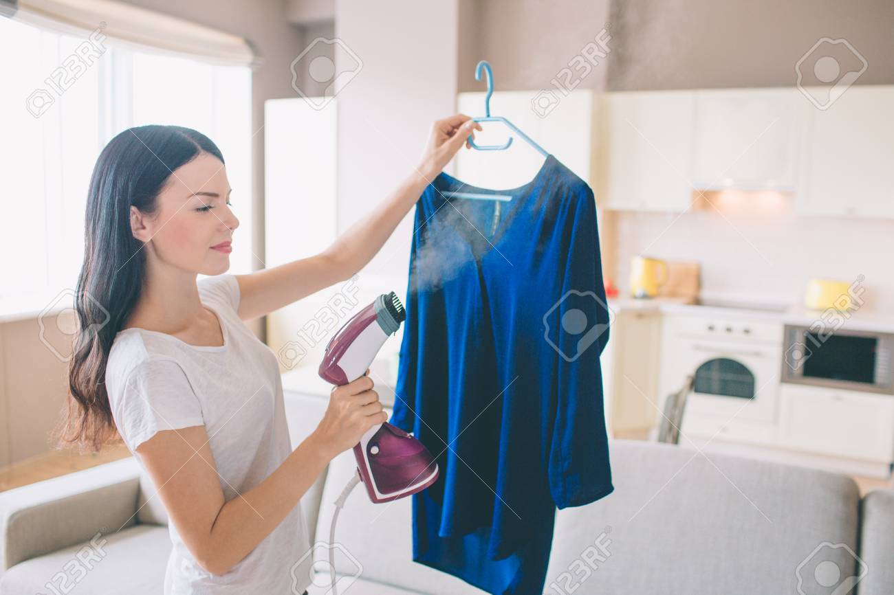 Woman is steaming blue shirt in room. She holds small stream iron in hand. Brunette is concentrated on work. - 108059929 Woman is steaming blue shirt in room. She holds small stream iron in hand. Brunette is concentrated on work. - 108059929