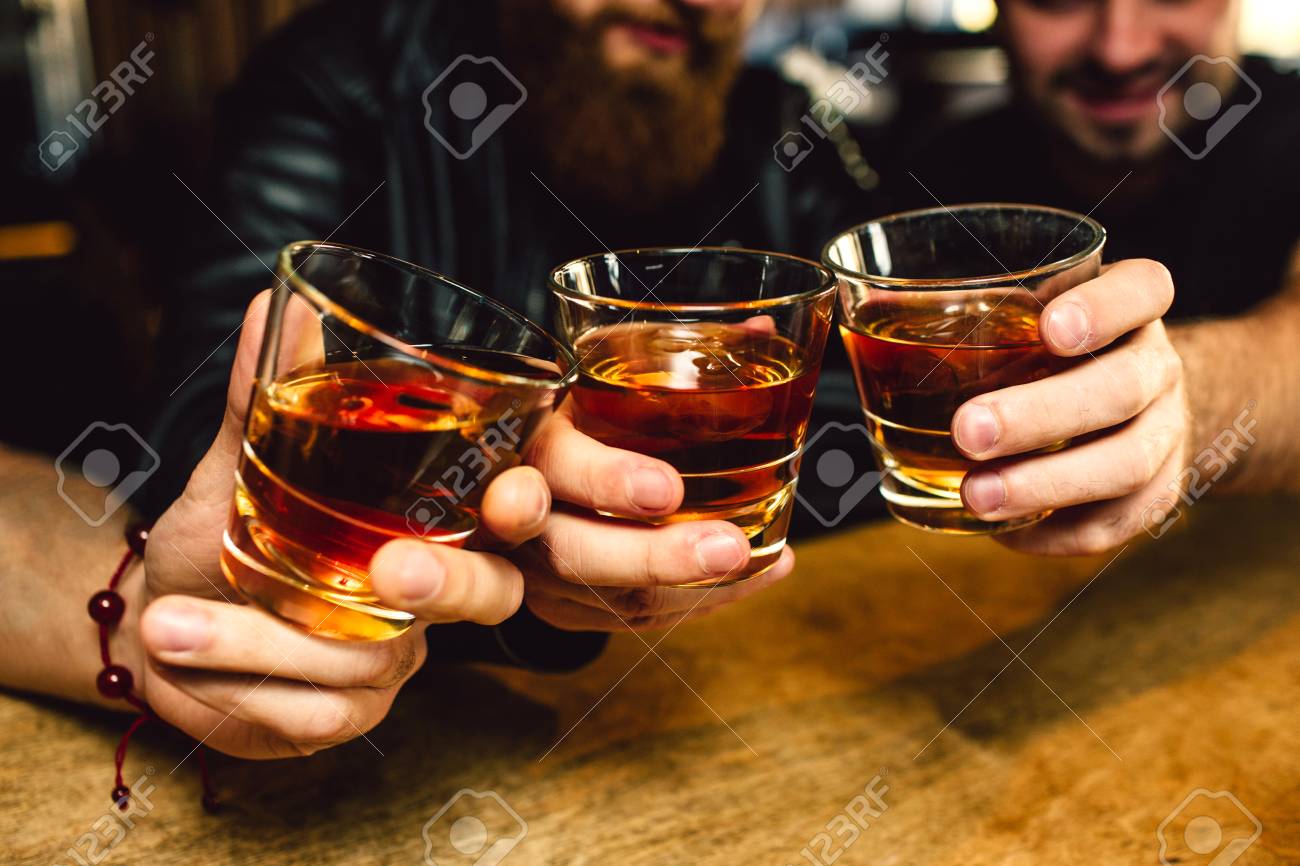 Cut view of three bearded young men holding glasses with rum together. They smile. People sit in bar - 119519705 Cut view of three bearded young men holding glasses with rum together. They smile. People sit in bar - 119519705