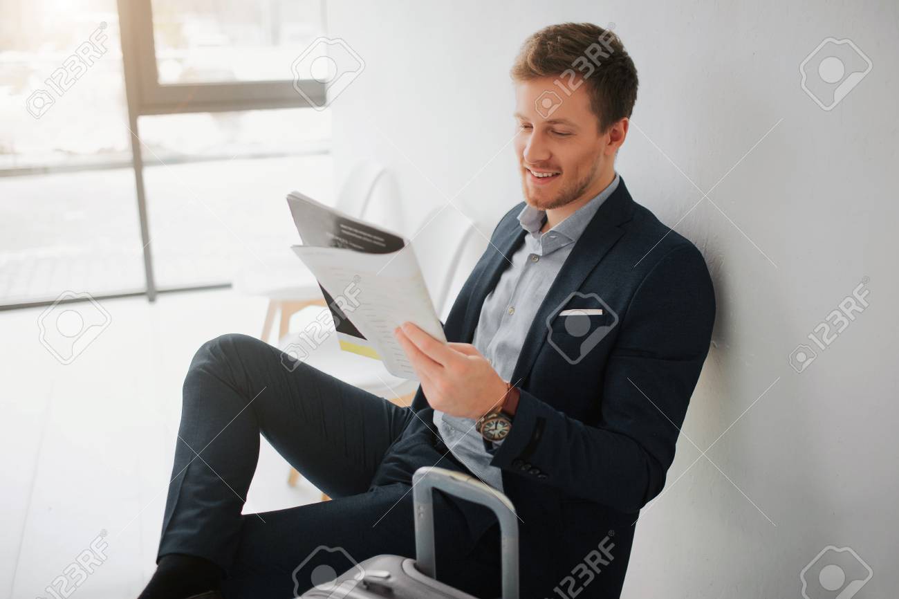 Cheerful young businessman sit on chair in white room. He read newspaper and smile. Guy wait for flight. - 115898175 Cheerful young businessman sit on chair in white room. He read newspaper and smile. Guy wait for flight. - 115898175