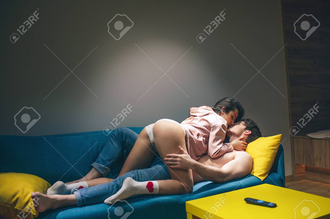 Young Couple Have In Kitchen In Night. Standing In Position On Sofa. Hot Seductive Woman On Top. Shirtless Man Lying On Sofa And Hold Hand On Her Hips. Sensual Moment