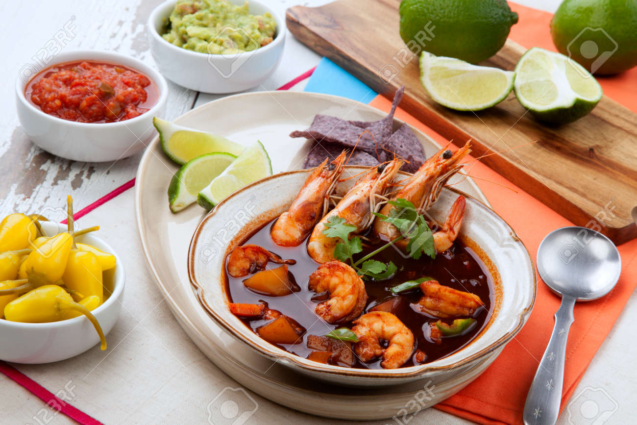 Closeup of a bowl of hot delicious spicy Mexican shrimp soup, garnished with whole shrimps and fresh cilantro. Limes, blue corn chips, salsa, guacamole, and marinated hot pepper. - 165024008 Closeup of a bowl of hot delicious spicy Mexican shrimp soup, garnished with whole shrimps and fresh cilantro. Limes, blue corn chips, salsa, guacamole, and marinated hot pepper. - 165024008