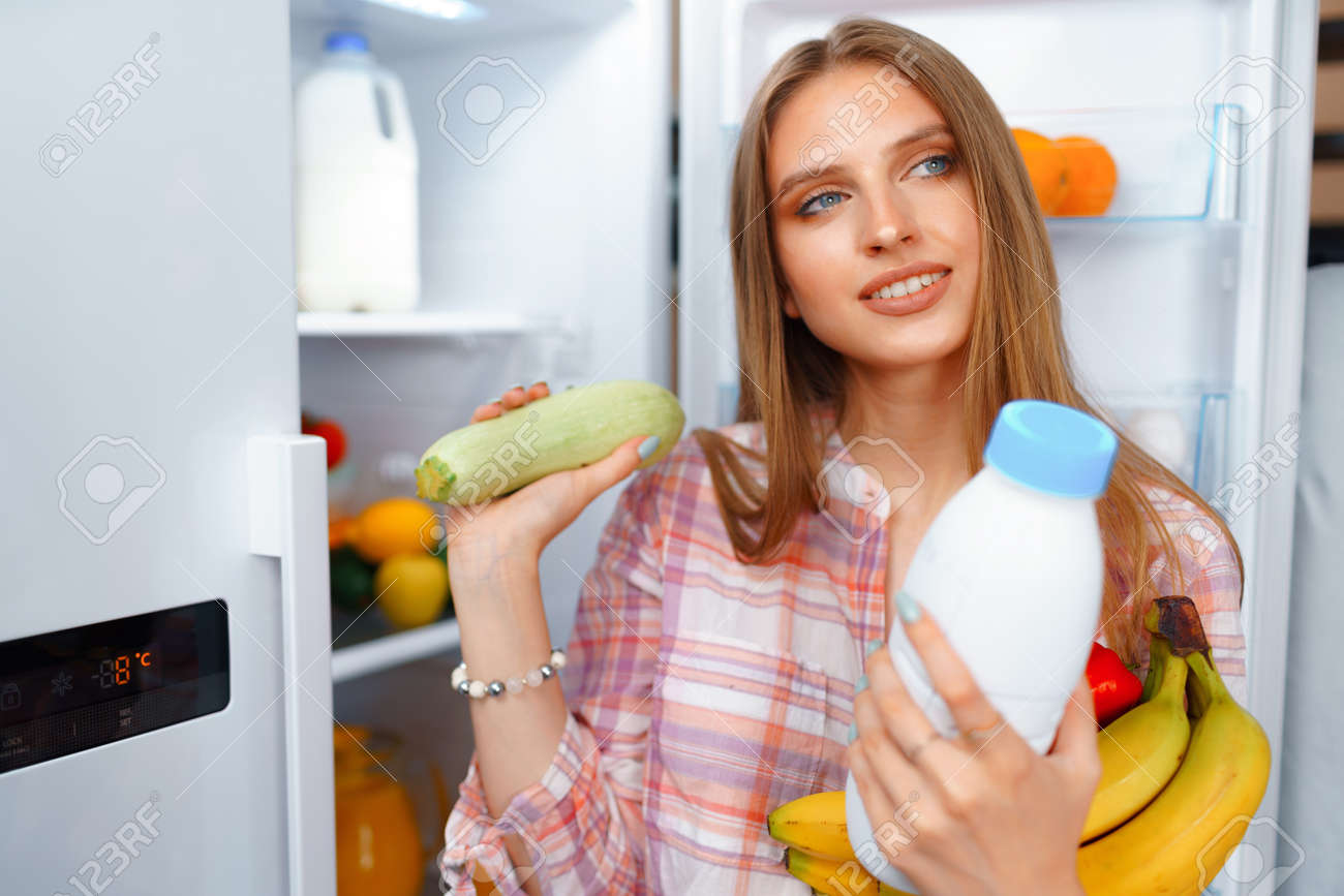 Portrait of a young blonde woman taking food from her fridge - 153483628 Portrait of a young blonde woman taking food from her fridge - 153483628