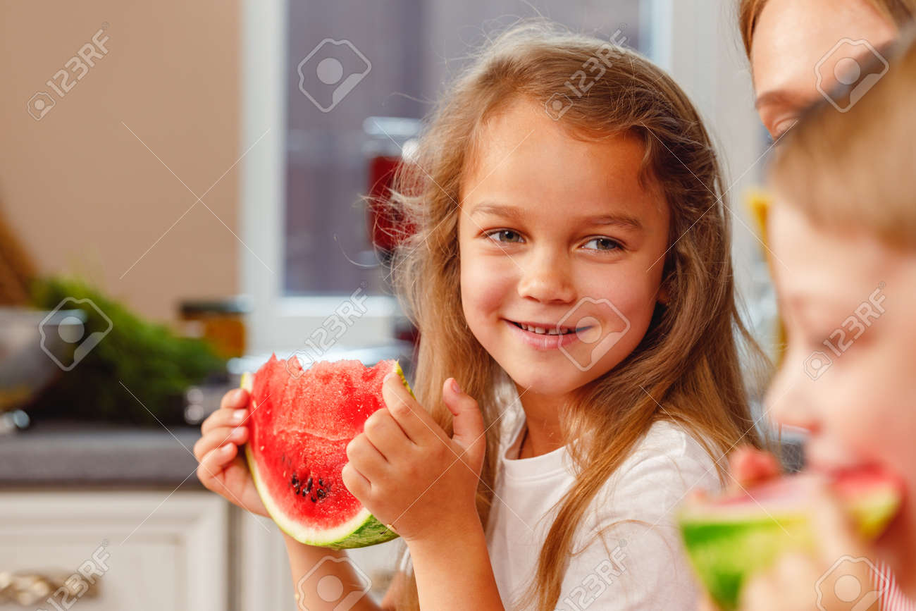 Smiling little girl eating a slice of watermelon in kitchen - 165810647 Smiling little girl eating a slice of watermelon in kitchen - 165810647