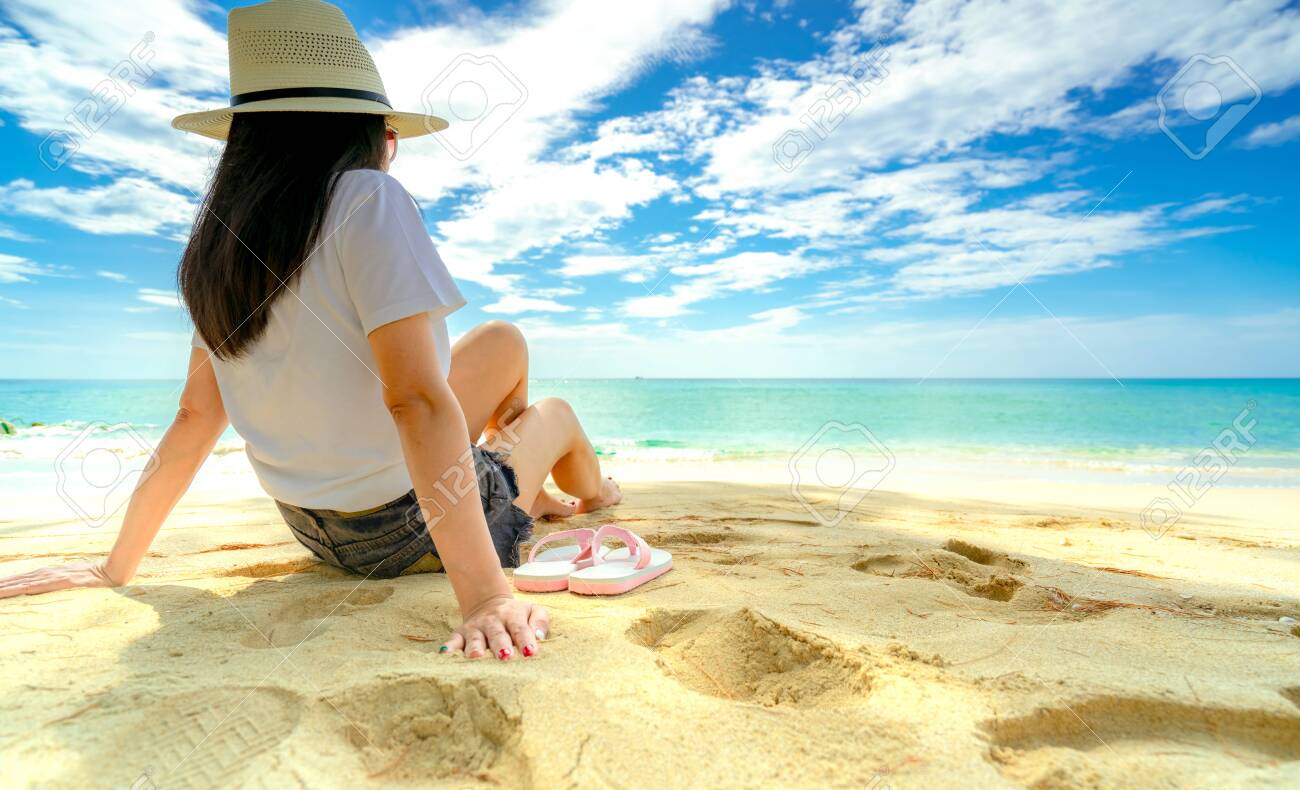 Happy Young Woman In White Shirts And Shorts Sitting At Sand Beach. Relaxing And Enjoying Holiday At Tropical Paradise Beach With Blue Sky And Clouds. Girl In Summer Vacation. Summer Vibes. Happy