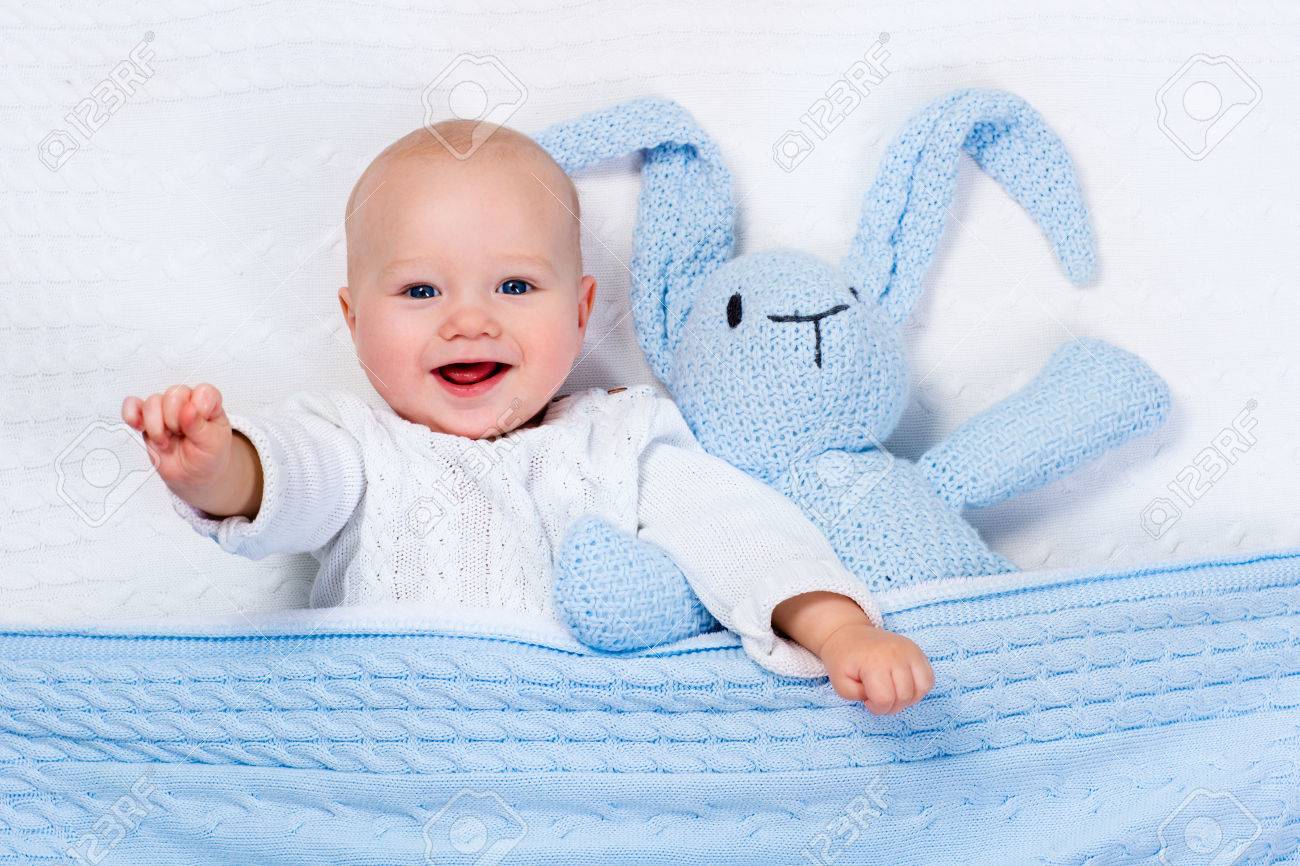 Funny little baby wearing a warm knitted jacket playing with toy bunny relaxing on white cable knit blanket in sunny nursery. Kids winter clothing and bedding. Hand made toys and textile for children. - 51358976 Funny little baby wearing a warm knitted jacket playing with toy bunny relaxing on white cable knit blanket in sunny nursery. Kids winter clothing and bedding. Hand made toys and textile for children. - 51358976