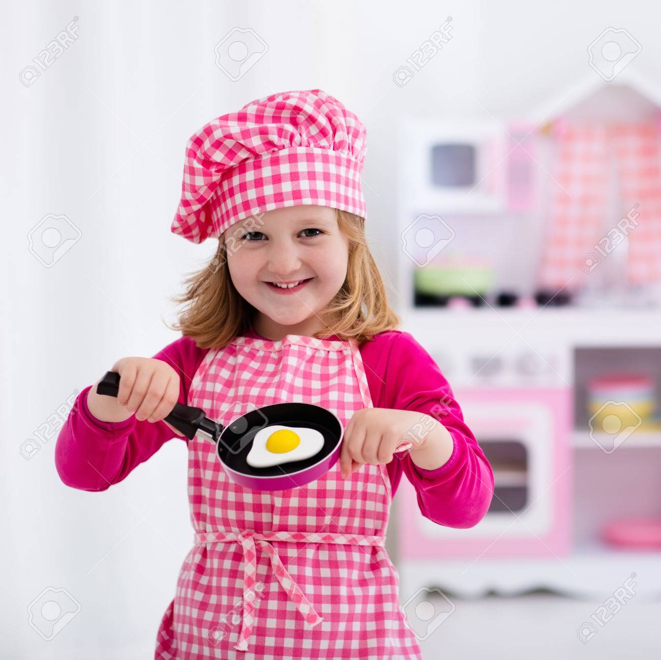Little Girl In Chef Hat And Apron Cooking Fried Eggs In Toy
