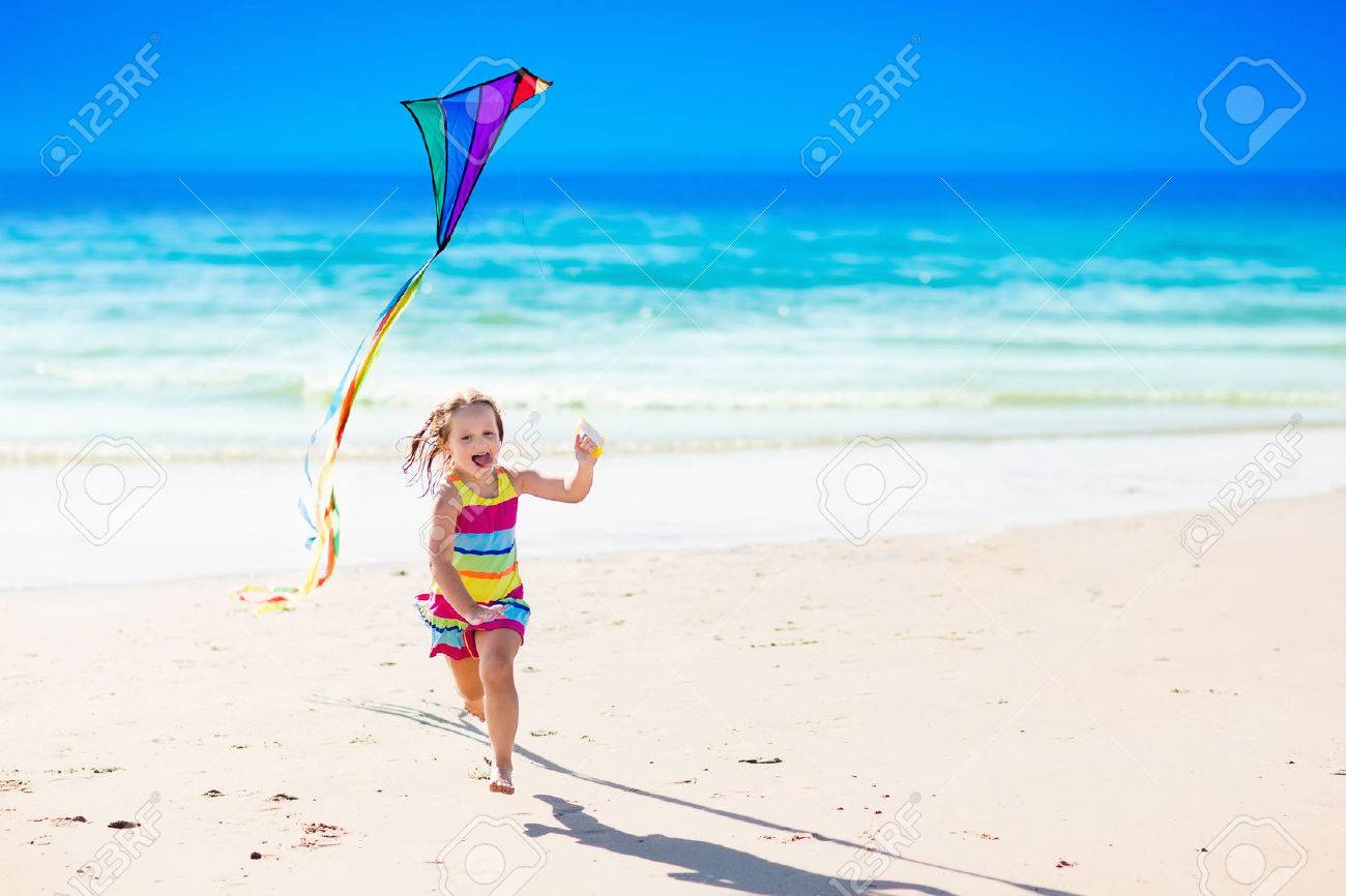 Happy laughing little girl flying a colorful kite running and jumping in sand on beautiful tropical beach during active summer family sea vacation. Kids play on ocean shore. Child with beach toys. - 75450337 Happy laughing little girl flying a colorful kite running and jumping in sand on beautiful tropical beach during active summer family sea vacation. Kids play on ocean shore. Child with beach toys. - 75450337