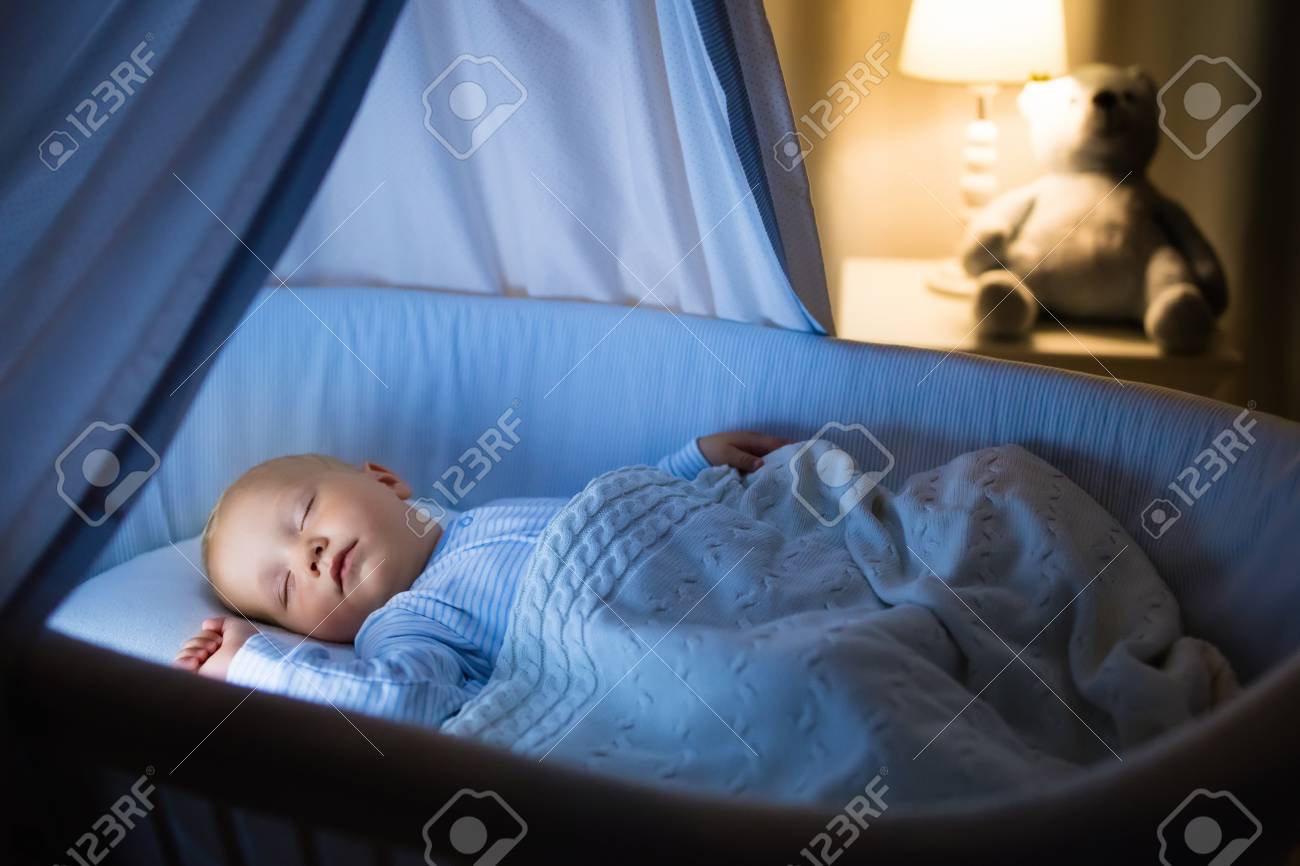 Adorable baby drinking milk in blue bassinet with canopy at night. Little boy in pajamas with formula bottle getting ready to sleep in dark room with crib, lamp and toy bear. Bed time drink for kids. - 87015351 Adorable baby drinking milk in blue bassinet with canopy at night. Little boy in pajamas with formula bottle getting ready to sleep in dark room with crib, lamp and toy bear. Bed time drink for kids. - 87015351