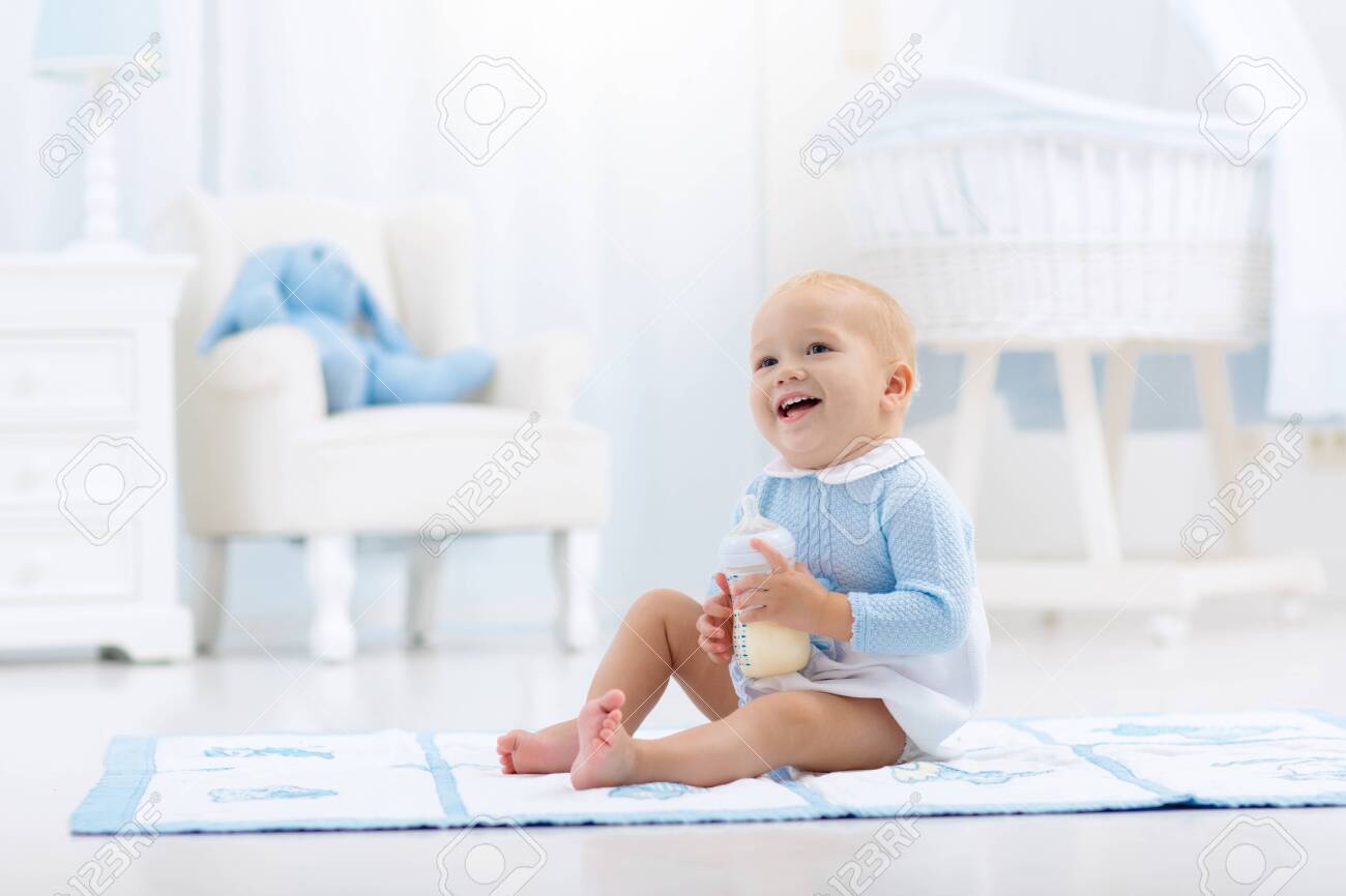 Adorable baby boy playing on a blue floor mat and drinking milk from a bottle in a white sunny nursery with rocking chair and bassinet. Bedroom interior with infant crib. Formula drink for infant. - 131756592 Adorable baby boy playing on a blue floor mat and drinking milk from a bottle in a white sunny nursery with rocking chair and bassinet. Bedroom interior with infant crib. Formula drink for infant. - 131756592