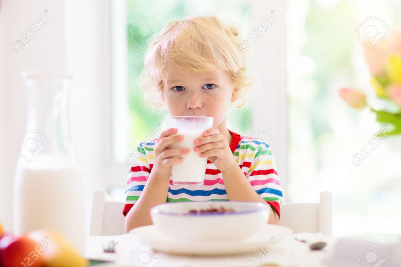 Child having breakfast. Kid drinking milk and eating cereal with fruit. Little boy at white dining table in kitchen at window. Kids eat on sunny morning. Healthy balanced nutrition for young kids. - 154589877 Child having breakfast. Kid drinking milk and eating cereal with fruit. Little boy at white dining table in kitchen at window. Kids eat on sunny morning. Healthy balanced nutrition for young kids. - 154589877