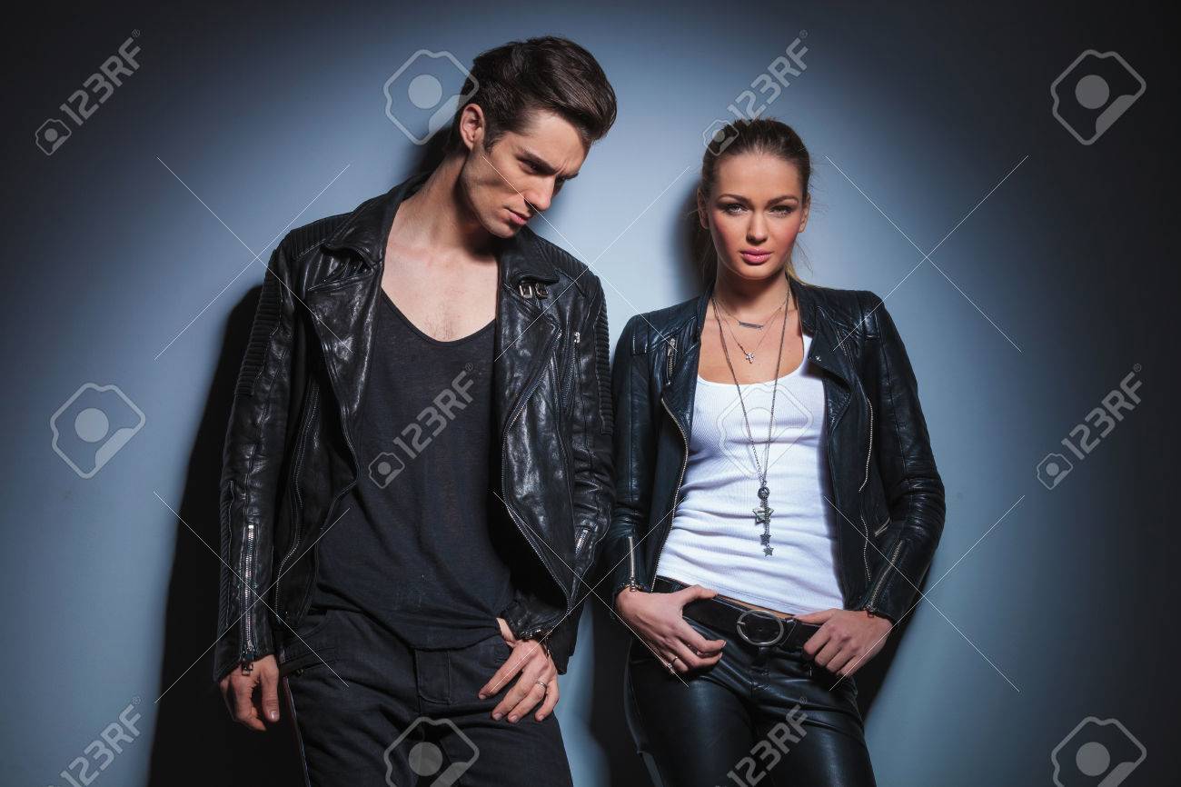 スタジオの背景で腰に手彼女の革のジャケットでポーズを取る彼のガール フレンド ポケットの手を見下ろして若いロッカー の写真素材 画像素材 Image