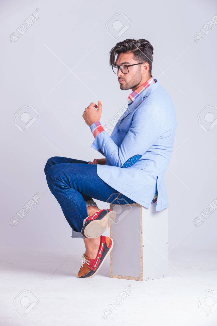 メガネを着用し スタジオの背景に目をそむけるながらあぐらをボックスの上に座って 1 つの側面からポーズ ファッショナブルなカジュアルな男 の写真素材 画像素材 Image