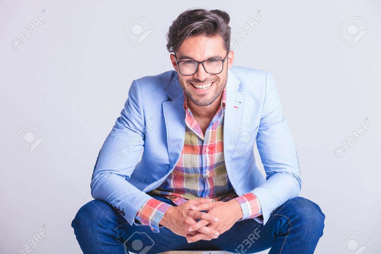 close portrait attractive man posing seated with legs spread open and hands touching, while smiling at the camera in studio background - 52312760 close portrait attractive man posing seated with legs spread open and hands touching, while smiling at the camera in studio background - 52312760