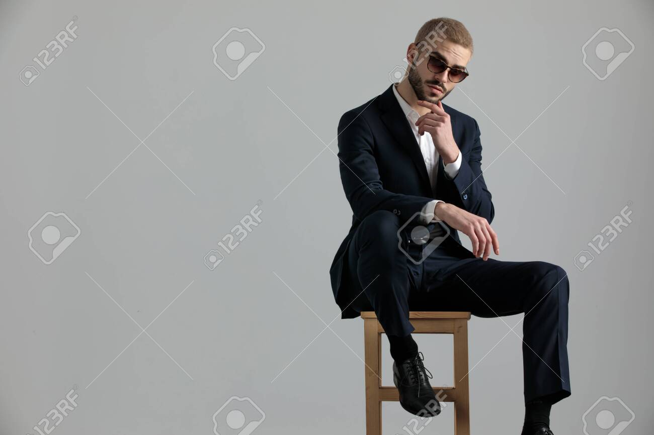 handsome formal business man wearing a navy suit and sunglasses sitting with one leg resting on a chair and touching his chin while looking at camera pensive on gray studio background - 130802330 handsome formal business man wearing a navy suit and sunglasses sitting with one leg resting on a chair and touching his chin while looking at camera pensive on gray studio background - 130802330