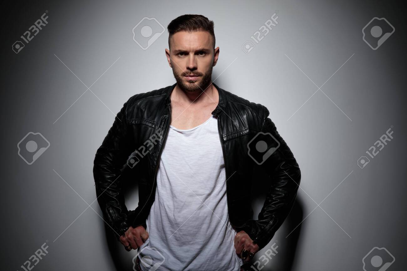 Tough young man adjusting his pants and looking forward while wearing a black leather jacket, standing on gray studio background - 133299719 Tough young man adjusting his pants and looking forward while wearing a black leather jacket, standing on gray studio background - 133299719