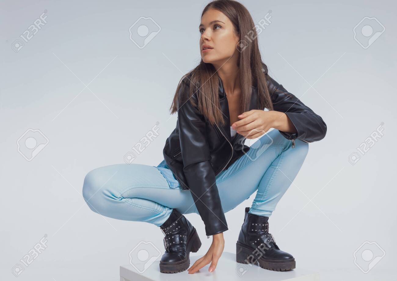 あおいれな しゃがみ 灰色のスタジオの背景にしゃがみながら革のジャケットを着て、目をそらしている焦点を当てたファッションモデルの写真素材・画像素材 Image 158048865