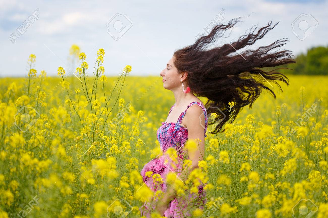 エスニック花柄のサンドレスに茶色の長い髪の毛との若い美しい女性は 髪を曇り空の背景で風になびかせ黄色の菜の花のフィールドに立っています の写真素材 画像素材 Image