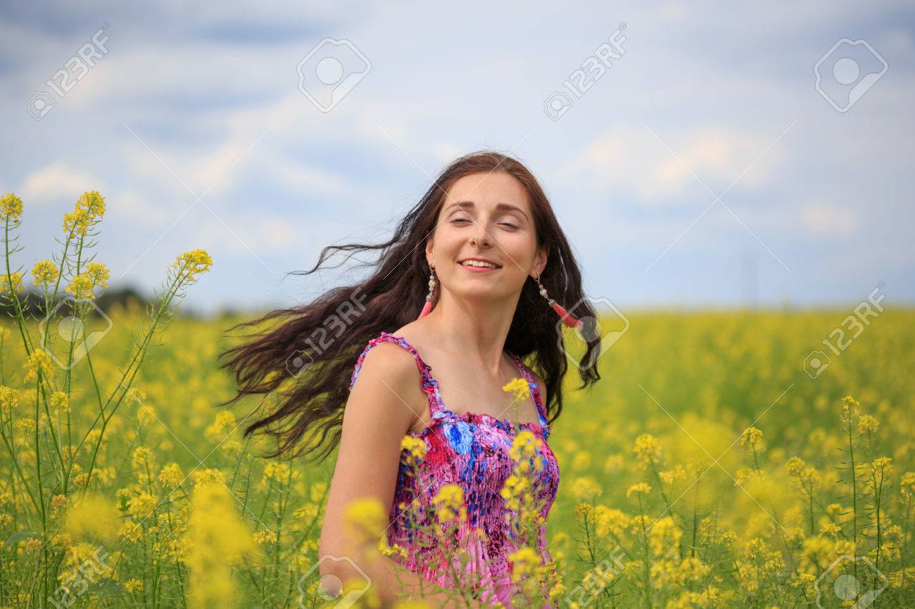 エスニック花柄のサンドレスに茶色の長い髪の毛との若い笑顔の女性は 髪を曇り空の背景で風になびかせ黄色の菜の花のフィールドに立っています の写真素材 画像素材 Image