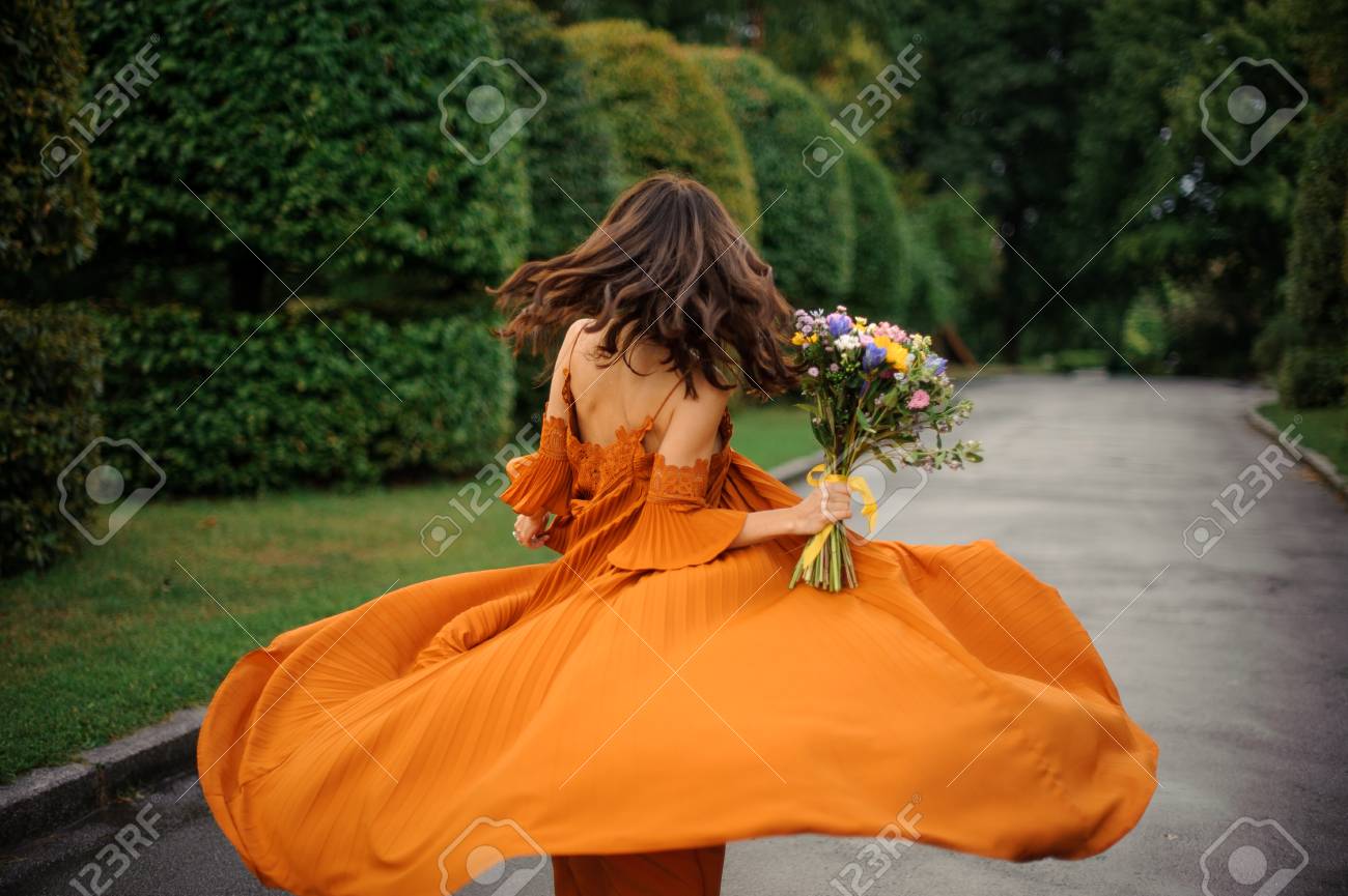 道路と木々 の背景に花の花束と長いオレンジのドレスで魅力的な女性の背面図 の写真素材 画像素材 Image