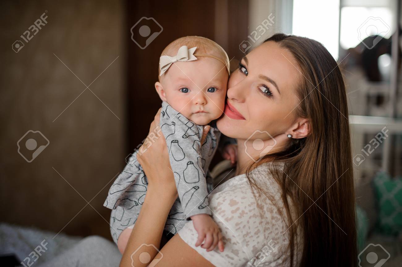 Stock Photo Young Brunette Mom Holding Her Little Blondie Daughter On Hands Close To Face In Their Home On The Blurred Background Stock Photo Young Brunette Mom Holding Her Little Blondie Daughter On Hands Close To Face In Their Home On The Blurred Background