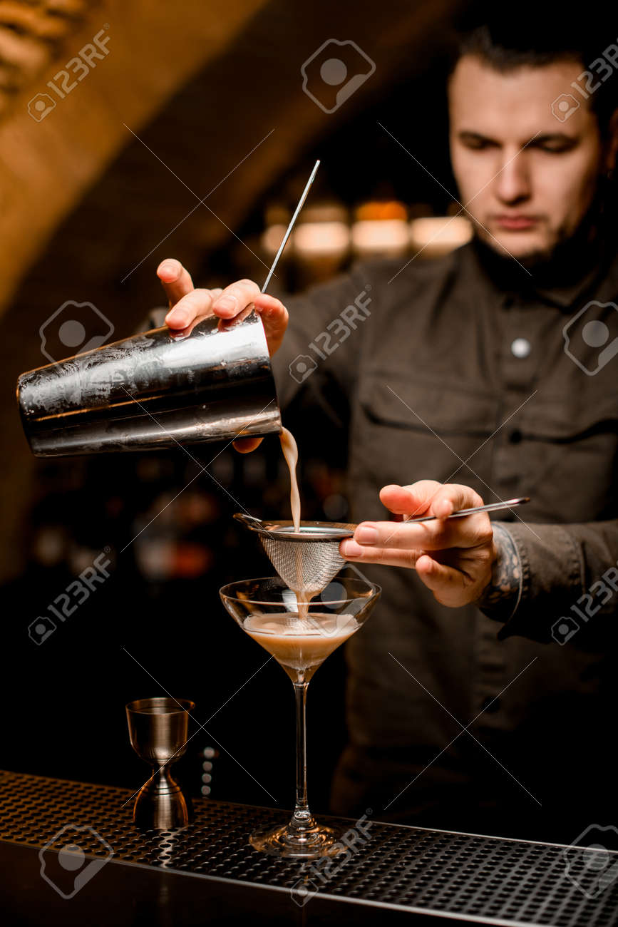 man bartender holds sieve and pours cocktail through it from shaker into glass - 168069117 man bartender holds sieve and pours cocktail through it from shaker into glass - 168069117