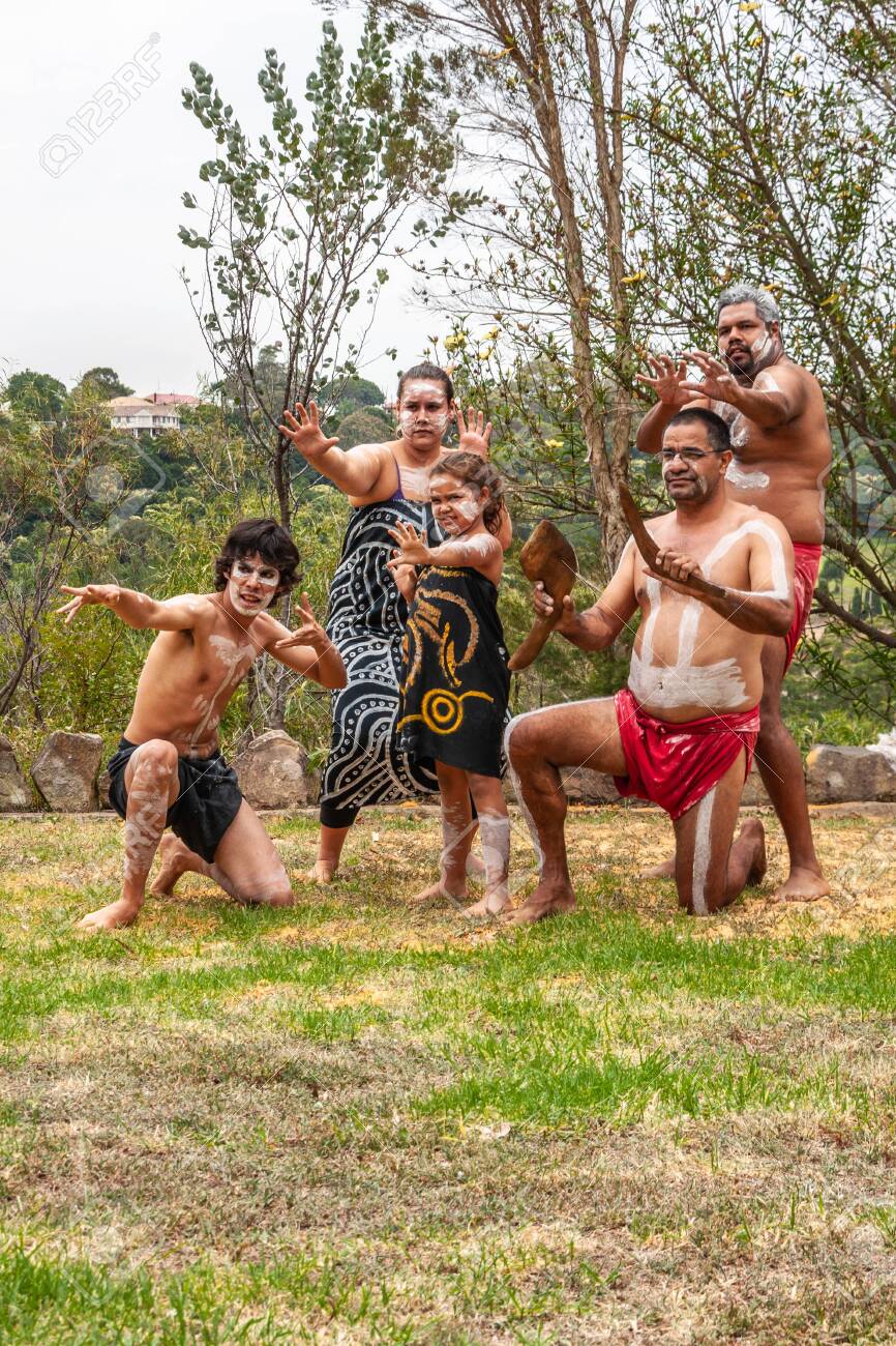 Newcastle, Australia - December 10, 2009: Aboriginal Family Strikes  Aggressive Pose As A Group On Green Grass With Green Foliage In Back. White  Stripes On Half-naked Bodies. Loincloth In Black And Red.