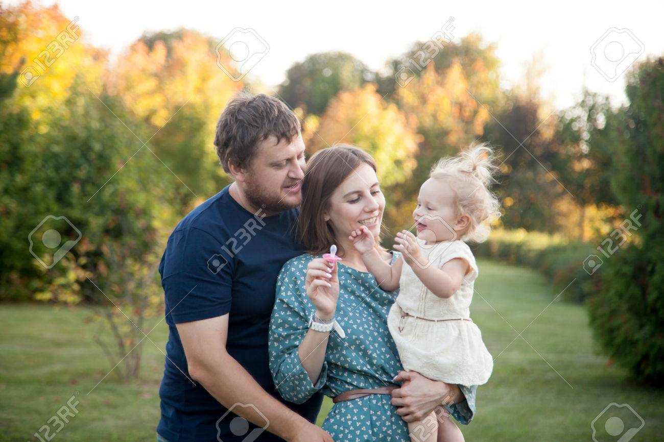 Retrato De Feliz Hermosa Familia De Tres Personas Caminando En El Parque En Otono Mama Y Papa Llevar A Su Hija Bebe En Vestido Blanco Jugando Tratando De Quitarle El Chupete Y