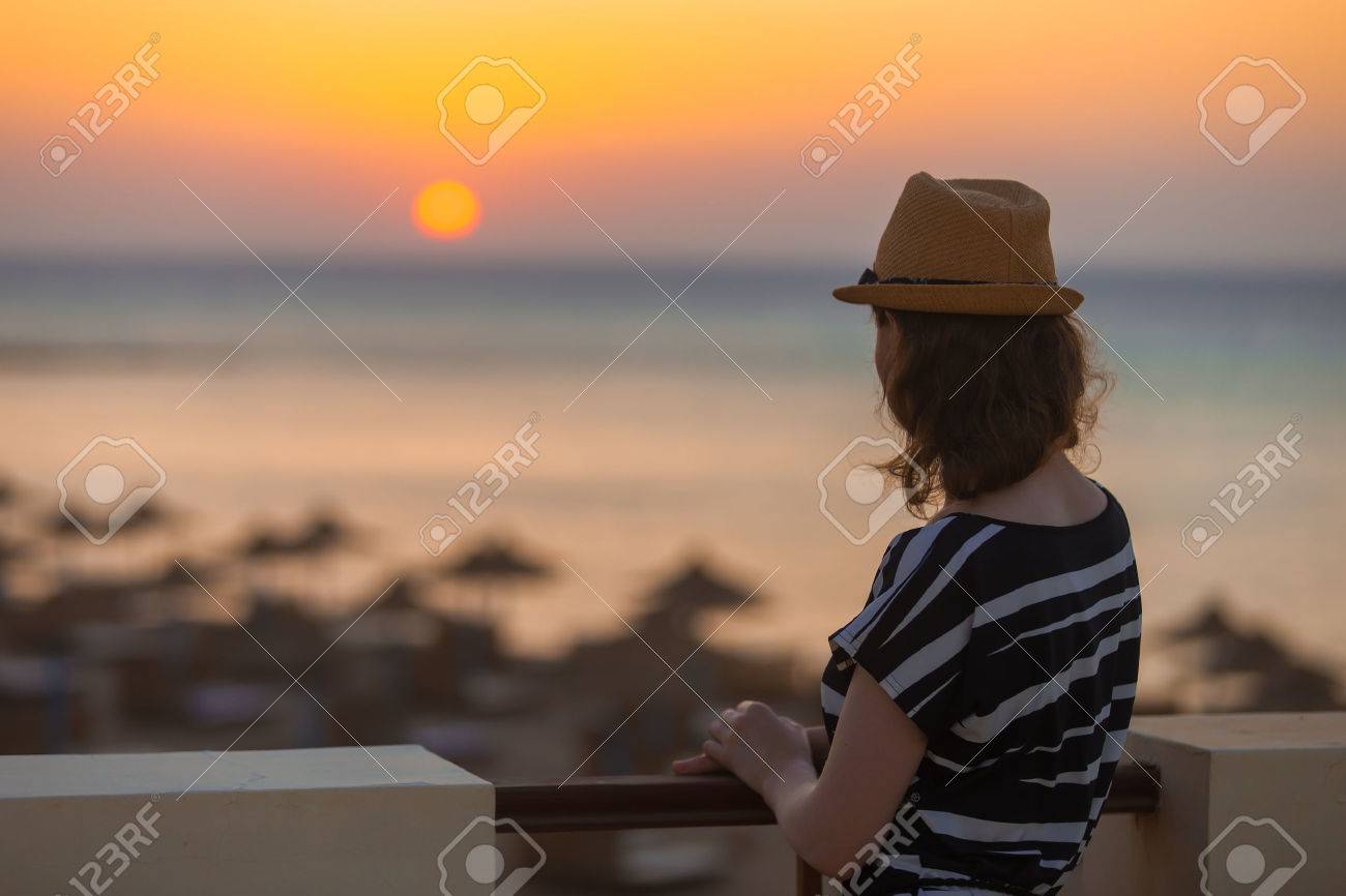 帽子とかわいい夏の若い女性地平線に夕日や日の出を見て 穏やかな海の風景を望むテラスに立ってのドレス 背面図 コピー スペース の写真素材 画像素材 Image