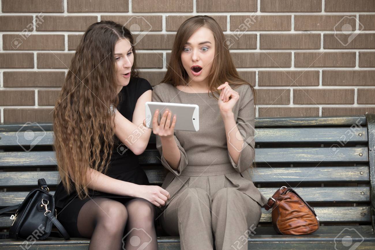 2 つの美しい若い驚いて恋人のベンチに座っているとショックを幸せそうな表情でタブレット画面を見ての肖像画 魅力的な女性の友達は有益な取り引きまたは販売 の写真素材 画像素材 Image