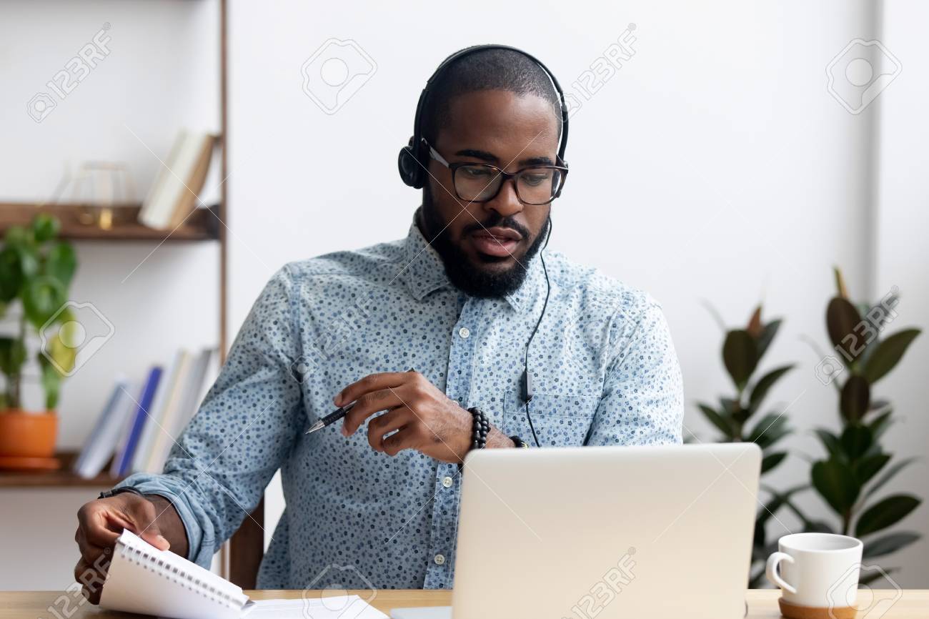 Black young man sitting at table wearing headphones learn foreign language improves knowledge looking at pc screen listening audio lesson holding pen and notepad makes some notes. E-learning concept - 114277925 Black young man sitting at table wearing headphones learn foreign language improves knowledge looking at pc screen listening audio lesson holding pen and notepad makes some notes. E-learning concept - 114277925