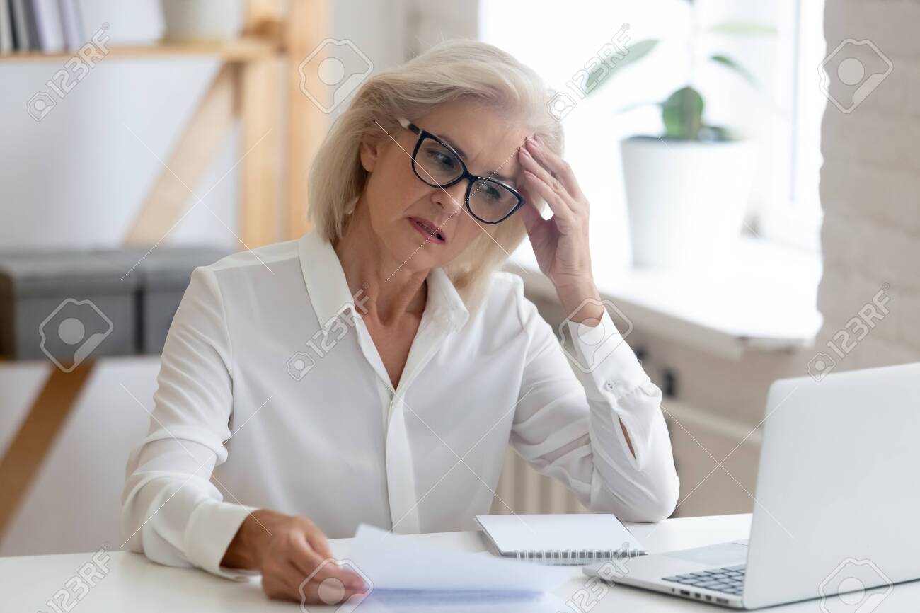 Pensive aged businesswoman in glasses sit at office desk thinking of problem solution, thoughtful senior woman worker in glasses look at laptop screen hold document pondering or making decision - 129607102 Pensive aged businesswoman in glasses sit at office desk thinking of problem solution, thoughtful senior woman worker in glasses look at laptop screen hold document pondering or making decision - 129607102