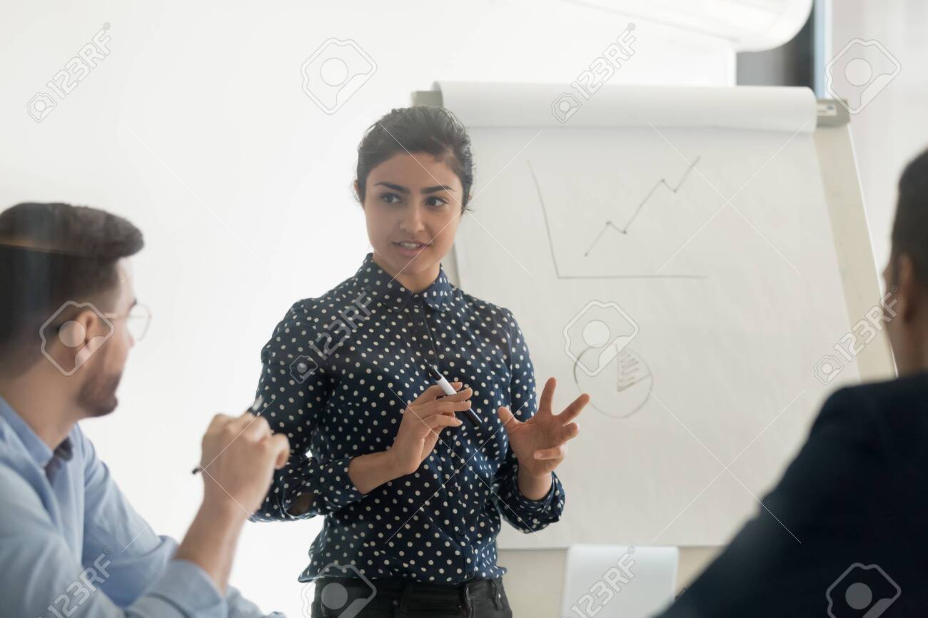Confident female indian conference speaker coach training diverse employees group give flip chart presentation at office meeting, hindu business woman presenter teach staff team at corporate workshop - 131244726 Confident female indian conference speaker coach training diverse employees group give flip chart presentation at office meeting, hindu business woman presenter teach staff team at corporate workshop - 131244726
