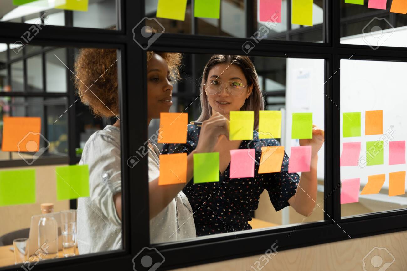 Concentrated african american young female scrum master working together with smart vietnamese businesswoman colleague, putting notes on sticky paper at window glass agile kanban board at office. - 133969051 Concentrated african american young female scrum master working together with smart vietnamese businesswoman colleague, putting notes on sticky paper at window glass agile kanban board at office. - 133969051