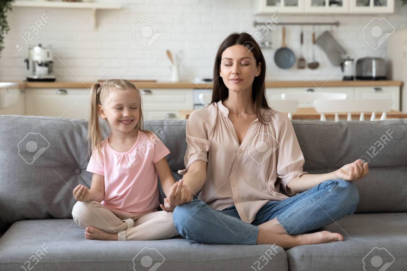 Front view mindful calm 30s woman sitting in lotus pose with cute little daughter on comfortable sofa in living room. Smiling preschool girl practicing yoga breathing exercises with mommy at home. - 133969161 Front view mindful calm 30s woman sitting in lotus pose with cute little daughter on comfortable sofa in living room. Smiling preschool girl practicing yoga breathing exercises with mommy at home. - 133969161