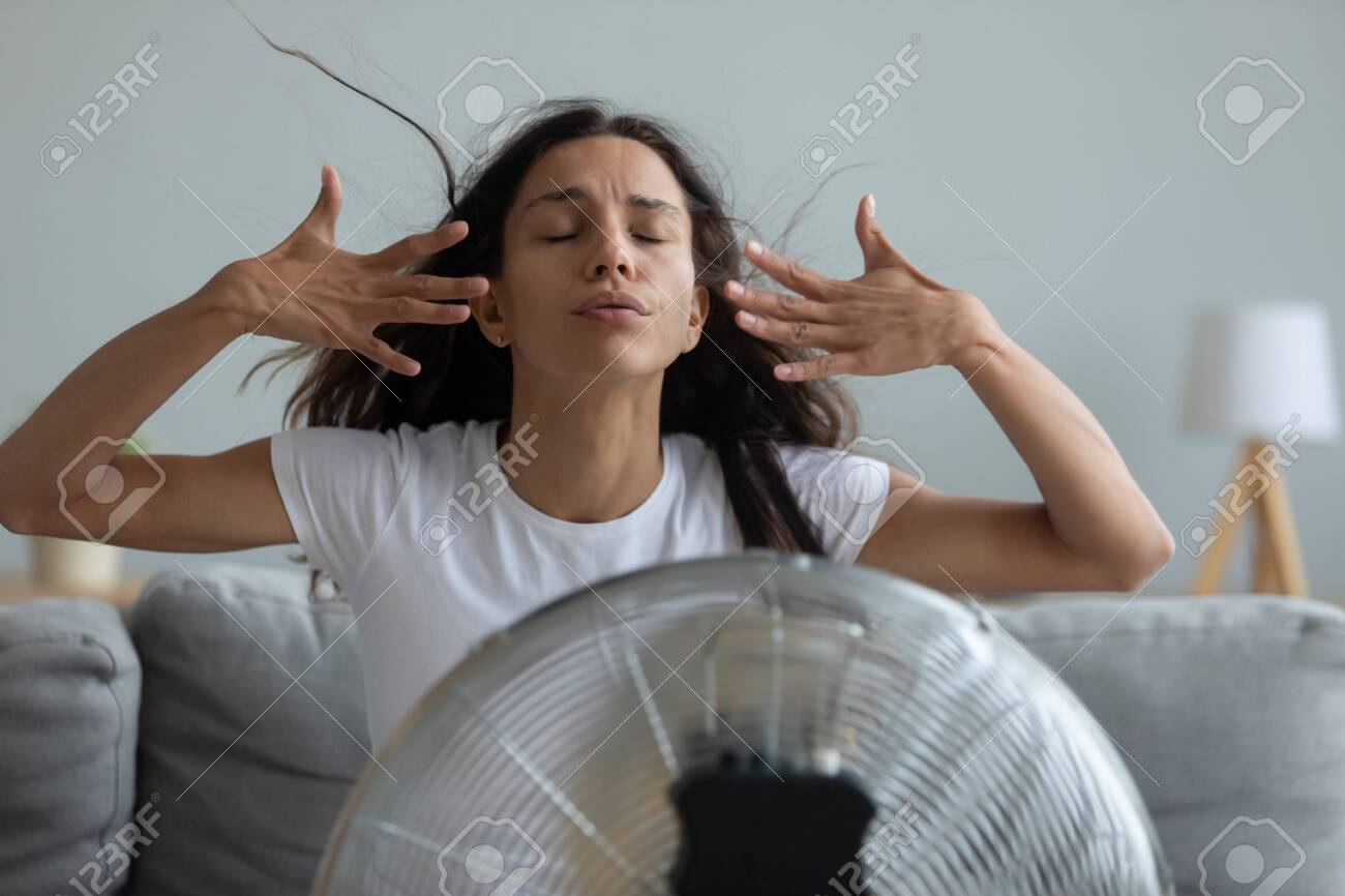 Woman seated on couch in living room suffers from swelter waving her hands trying to cool off by the fan, oppressive heat too hot weather and sultriness in summer, flat without air-conditioner concept - 134207753 Woman seated on couch in living room suffers from swelter waving her hands trying to cool off by the fan, oppressive heat too hot weather and sultriness in summer, flat without air-conditioner concept - 134207753