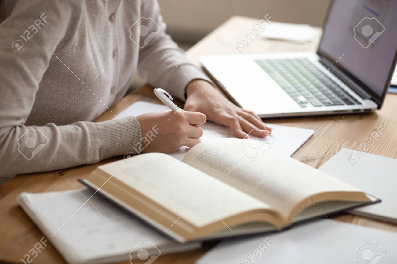 Close up female student sitting at desk, writing down notes from opened textbook and laptop. Young girl preparing for university session, exams, doing homework, paperwork, writing coursework or essay. - 134439496 Close up female student sitting at desk, writing down notes from opened textbook and laptop. Young girl preparing for university session, exams, doing homework, paperwork, writing coursework or essay. - 134439496