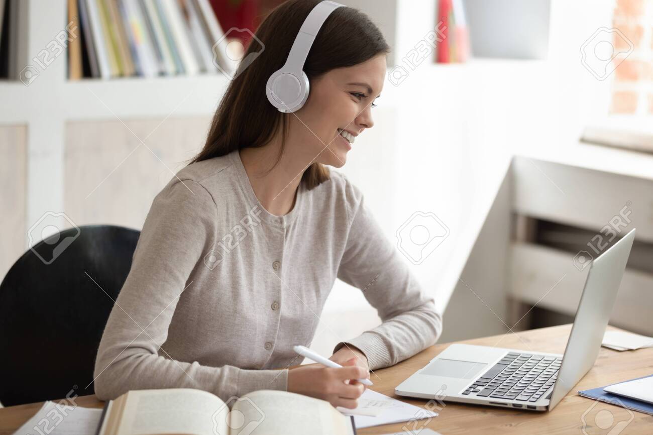 Smiling young female student wearing headset, looking at computer screen, listening interesting online lecture or watching educational video, writing down notes, doing homework or preparing for exam. - 134439577 Smiling young female student wearing headset, looking at computer screen, listening interesting online lecture or watching educational video, writing down notes, doing homework or preparing for exam. - 134439577