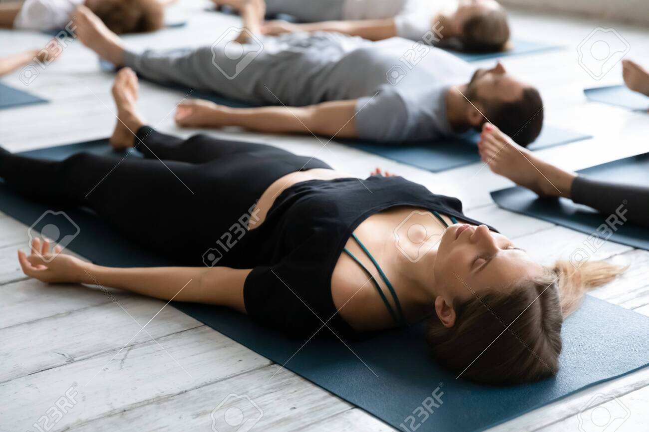 Beautiful young woman meditating in Savasana pose on floor close up, practicing yoga at group lesson, doing Corpse exercise on mats, training, working out in modern yoga studio, center - 138134929 Beautiful young woman meditating in Savasana pose on floor close up, practicing yoga at group lesson, doing Corpse exercise on mats, training, working out in modern yoga studio, center - 138134929
