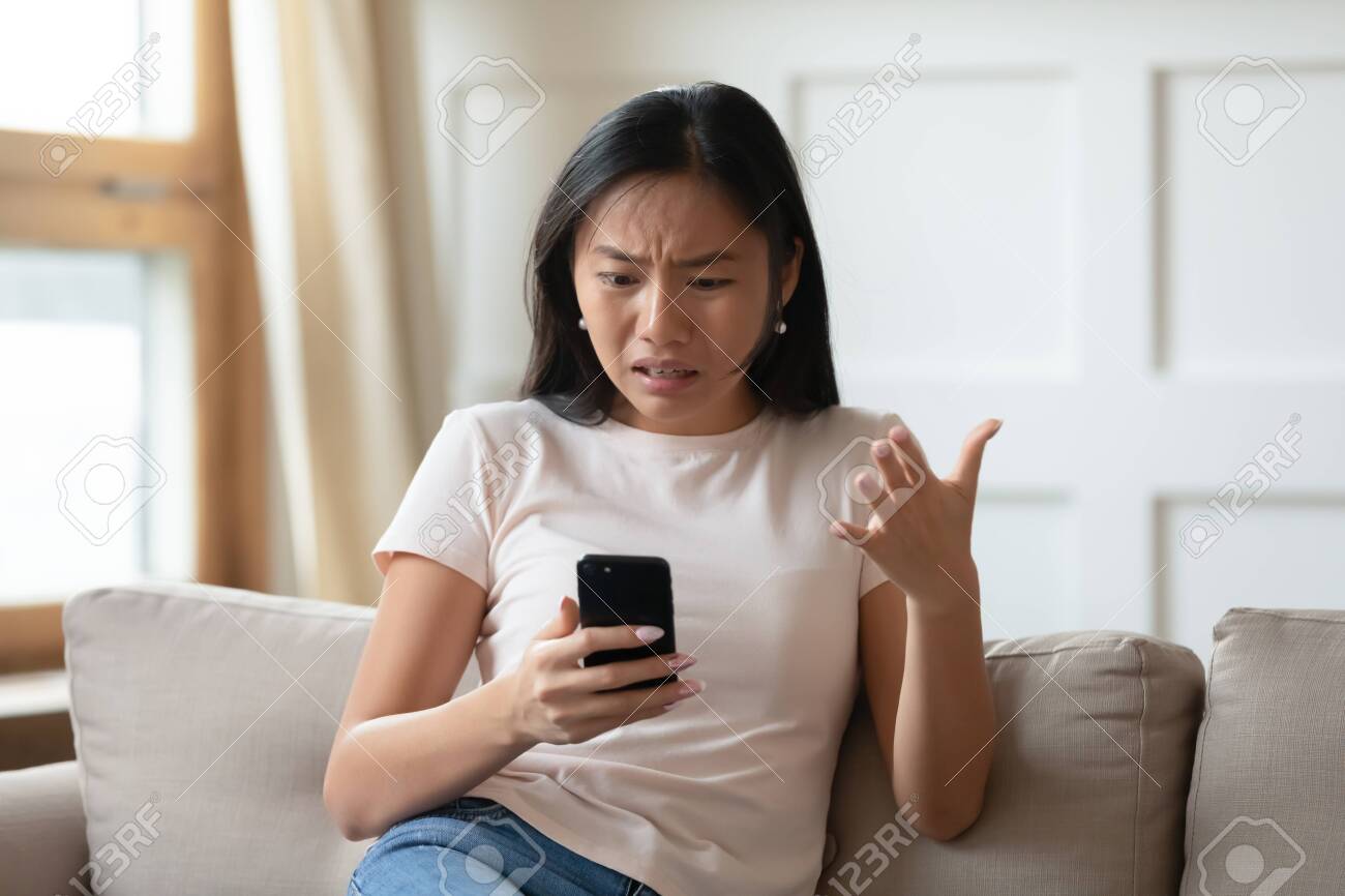 Annoyed Asian millennial girl sit on couch in living room frustrated with slow Internet connection or spam on smartphone, mad Vietnamese young woman confused with cellphone device operational problem - 138990269 Annoyed Asian millennial girl sit on couch in living room frustrated with slow Internet connection or spam on smartphone, mad Vietnamese young woman confused with cellphone device operational problem - 138990269