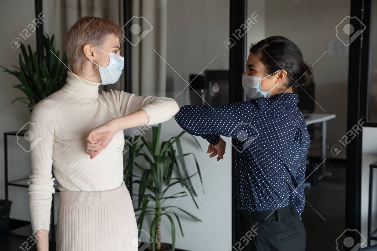 Smiling diverse female colleagues wearing protective face masks greeting bumping elbows at workplace, happy coworkers in medical facial covers protect from COVID-19 pandemics, healthcare concept - 145351859 Smiling diverse female colleagues wearing protective face masks greeting bumping elbows at workplace, happy coworkers in medical facial covers protect from COVID-19 pandemics, healthcare concept - 145351859