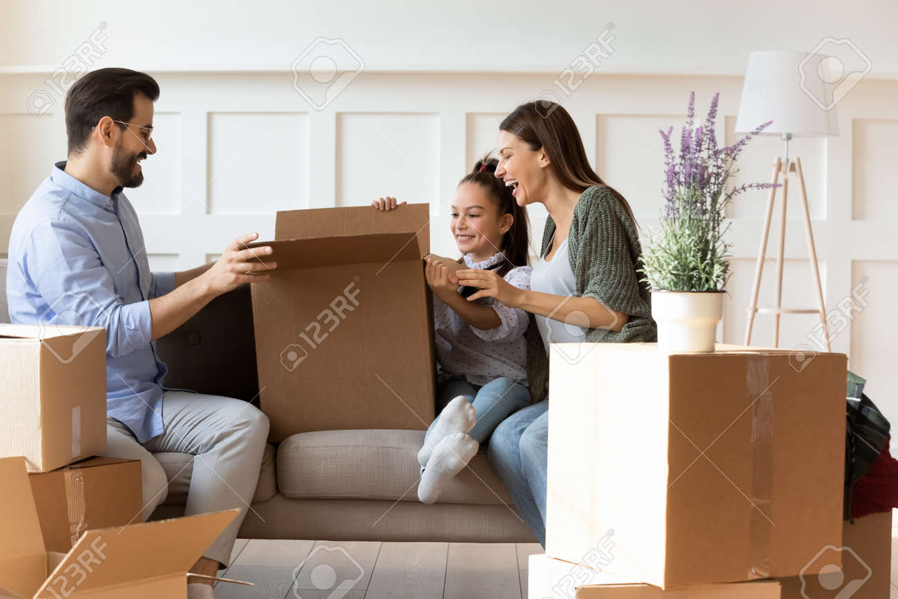 Adorable daughter helping parents with cardboard boxes on moving day, happy family with child sitting on couch, unpacking belongings in modern living room, relocation and mortgage concept - 149841651 Adorable daughter helping parents with cardboard boxes on moving day, happy family with child sitting on couch, unpacking belongings in modern living room, relocation and mortgage concept - 149841651